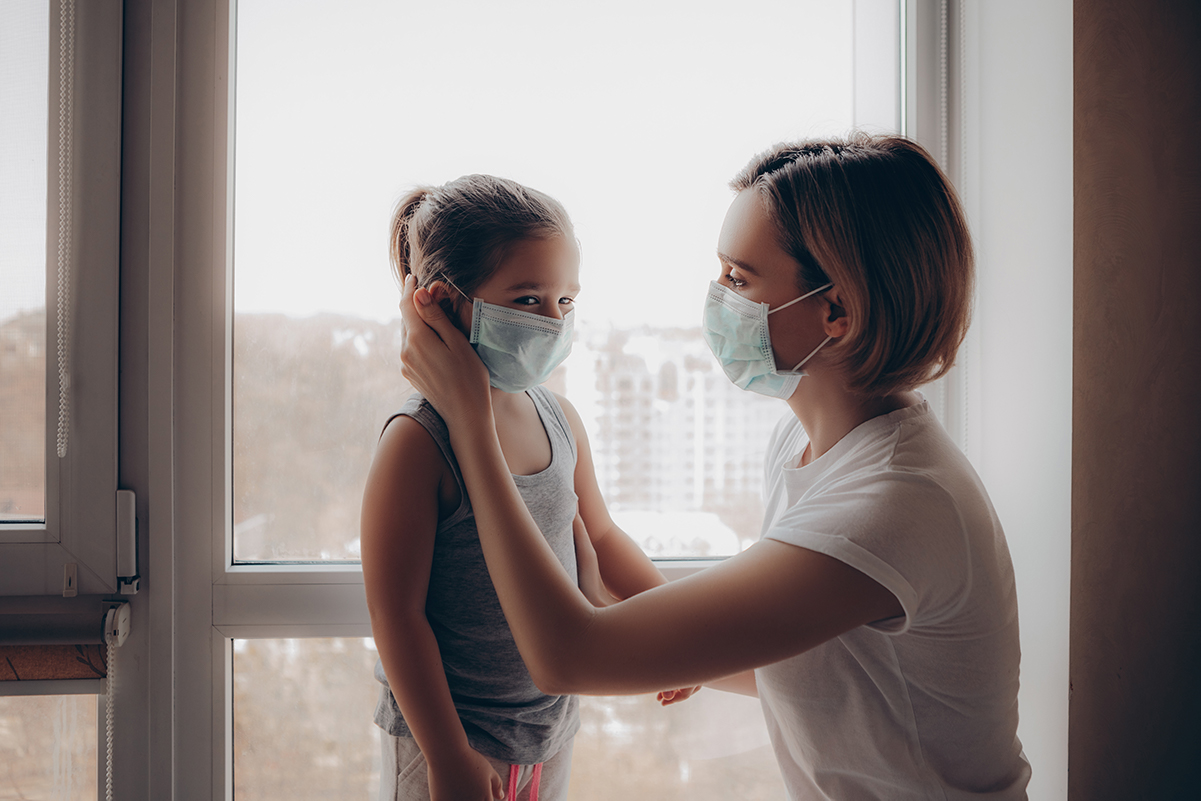 Mom putting mask on daughter standing in front of a window