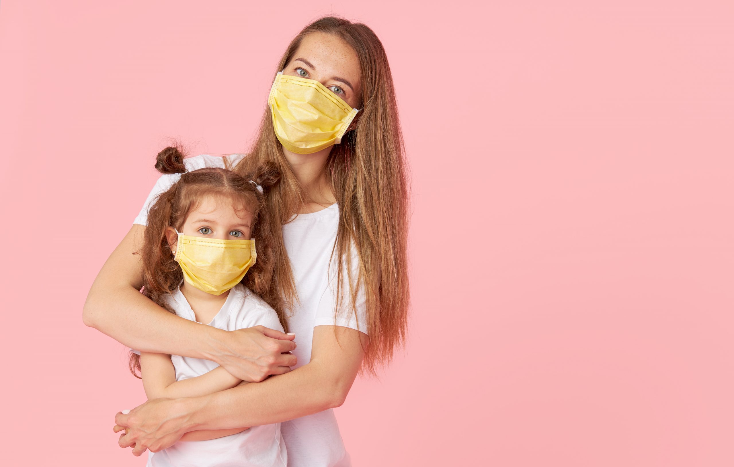 Caring for loved ones. Young mother hugs little daughter in protective medical masks during Covid-19 pandemics. Studio pink background