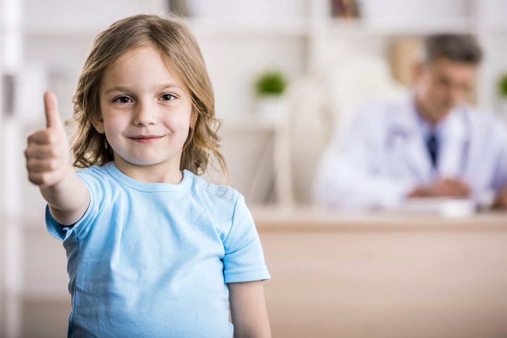 Cute little girl is showing thumb up. Doctor is sitting on background.