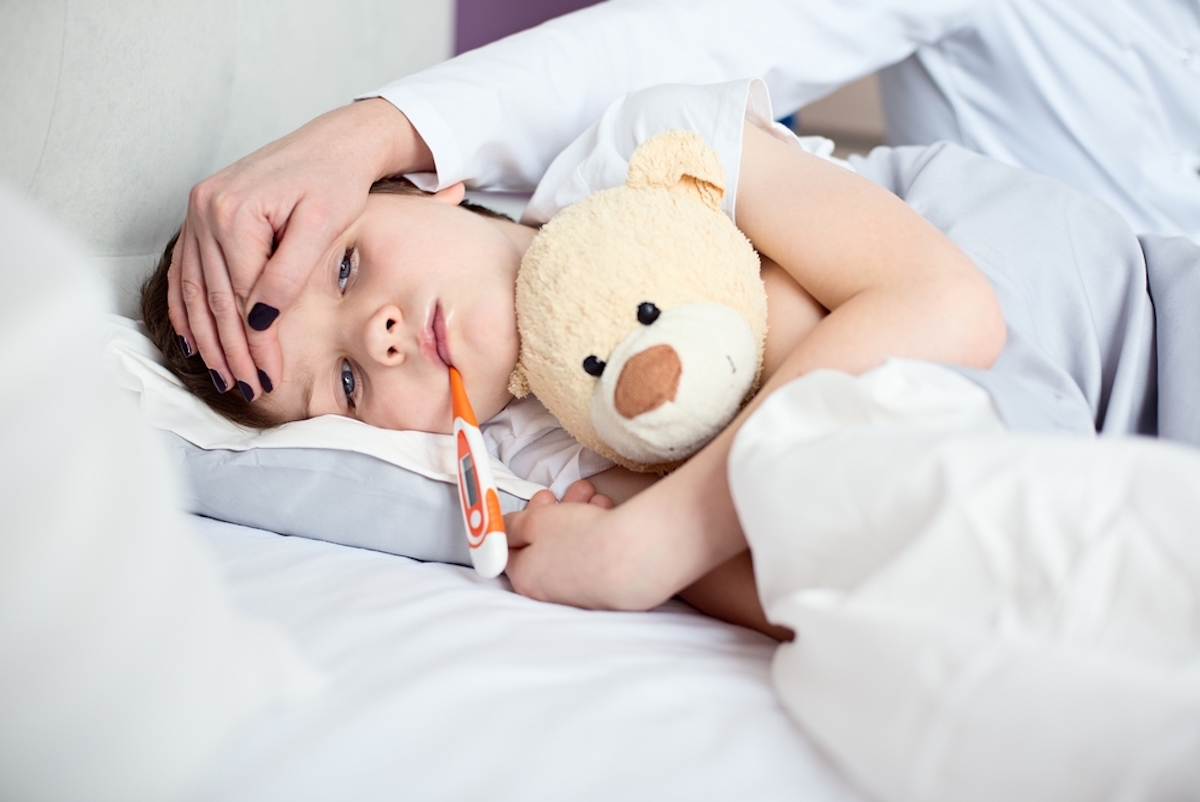 young boy sick with flu with thermometer in his mouth and teddy bear in his arms