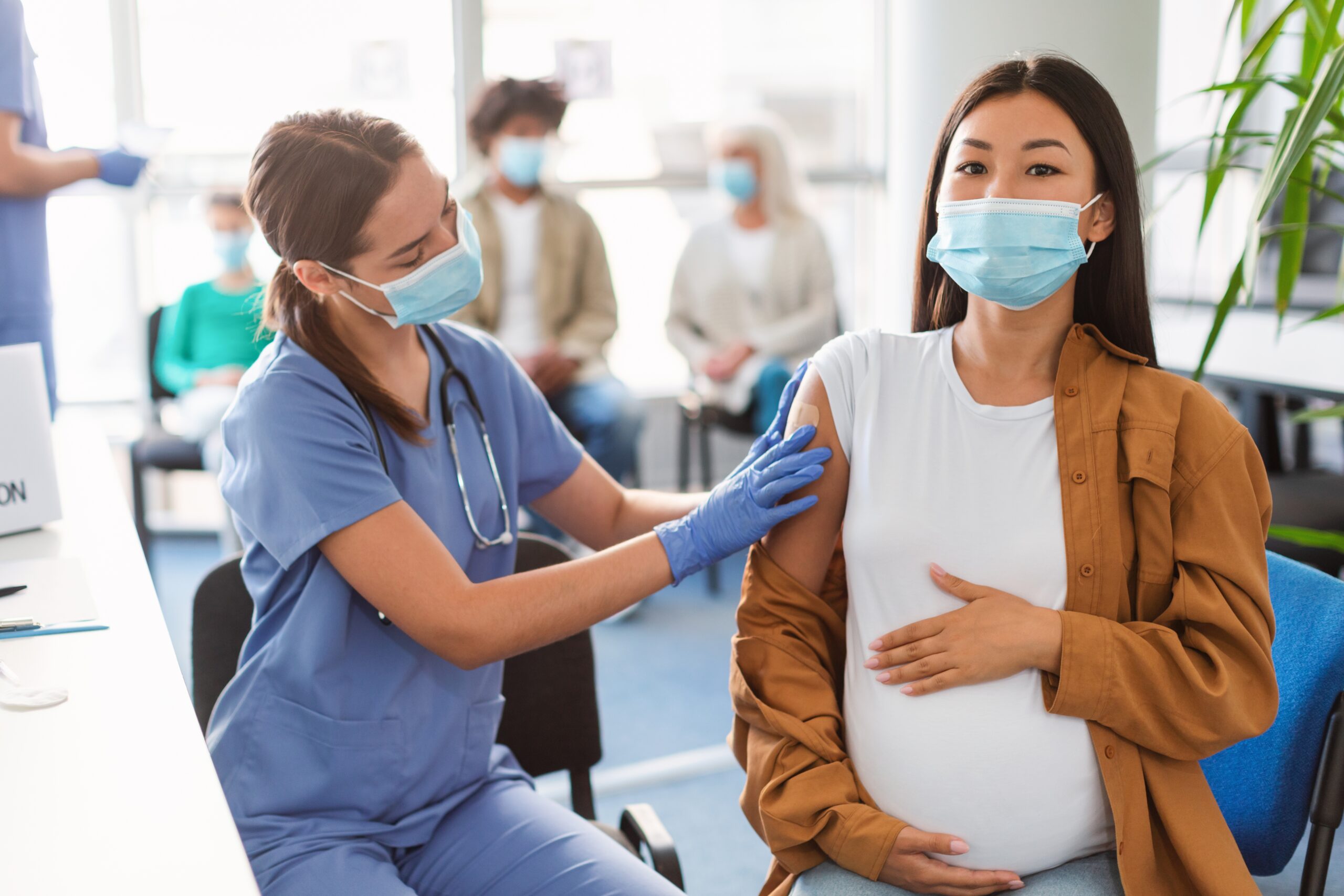 Asian pregnant woman wearing a mask is getting a bandage placed on her arm by a white female nurse in blue scrubs and a mask.