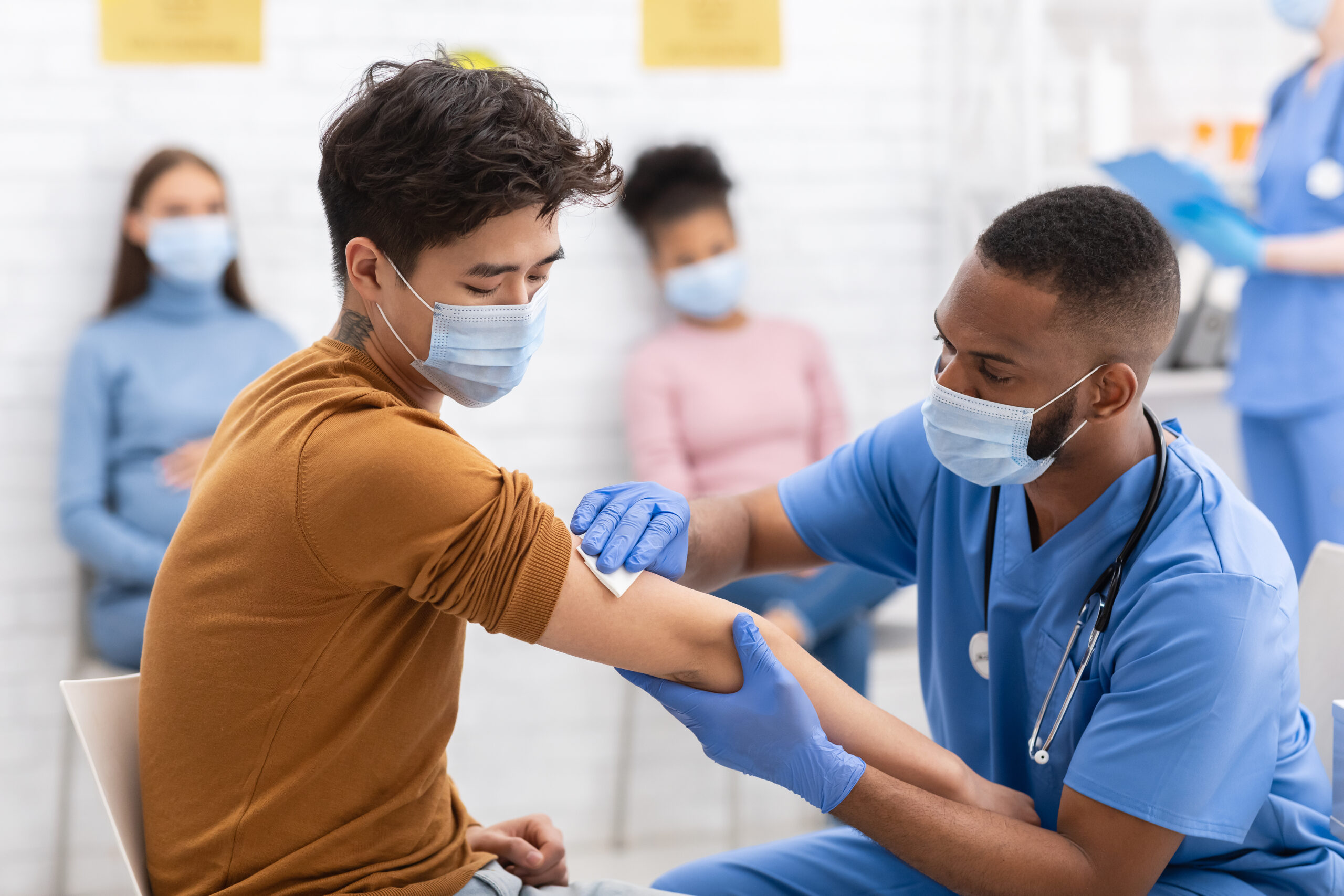 Asian man receives bandage on arm from an African American male nurse in blue scrubs. Both wearing masks