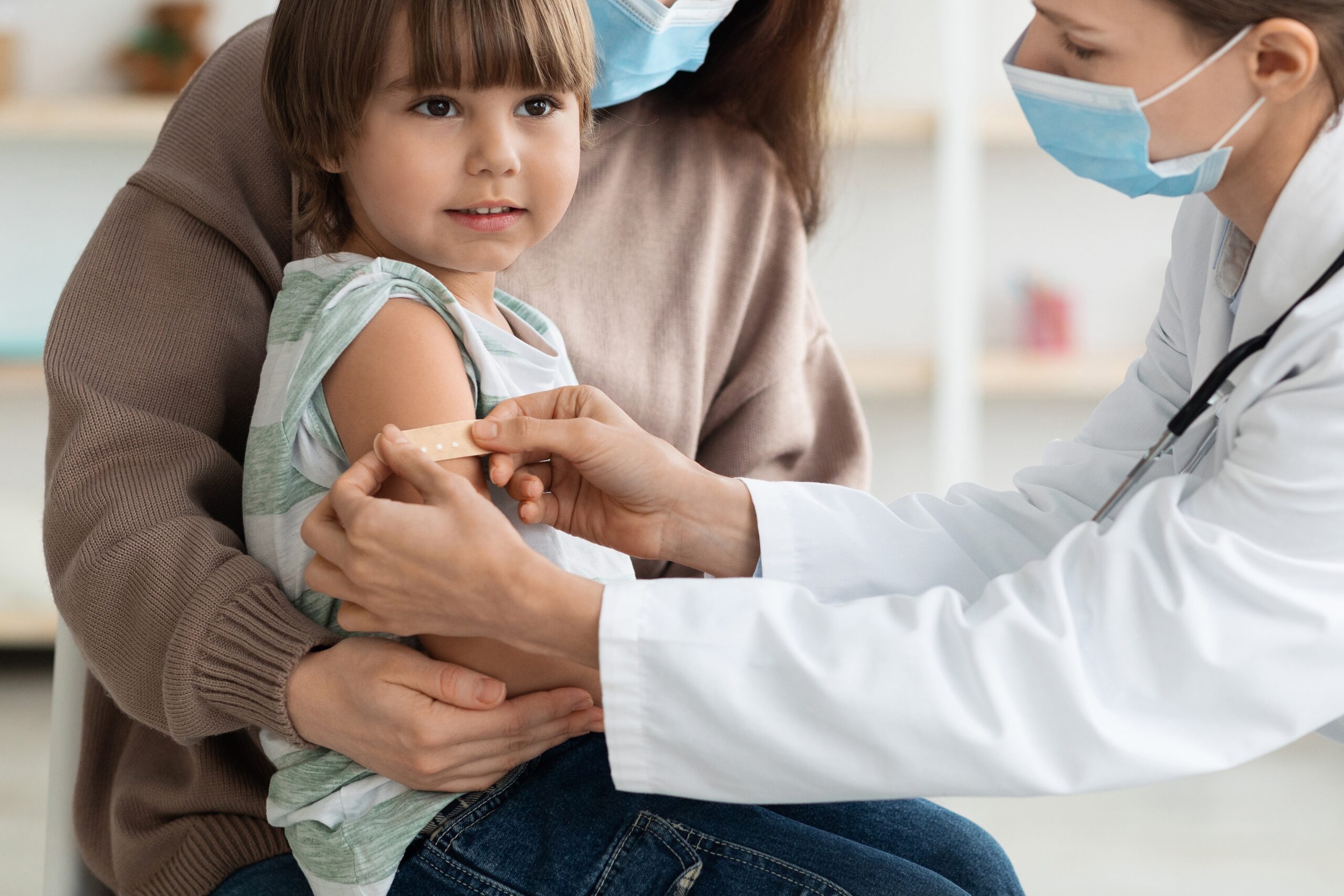 Vaccinated child getting a bandaid put on her arm by a doctor while sitting on her mom's lap