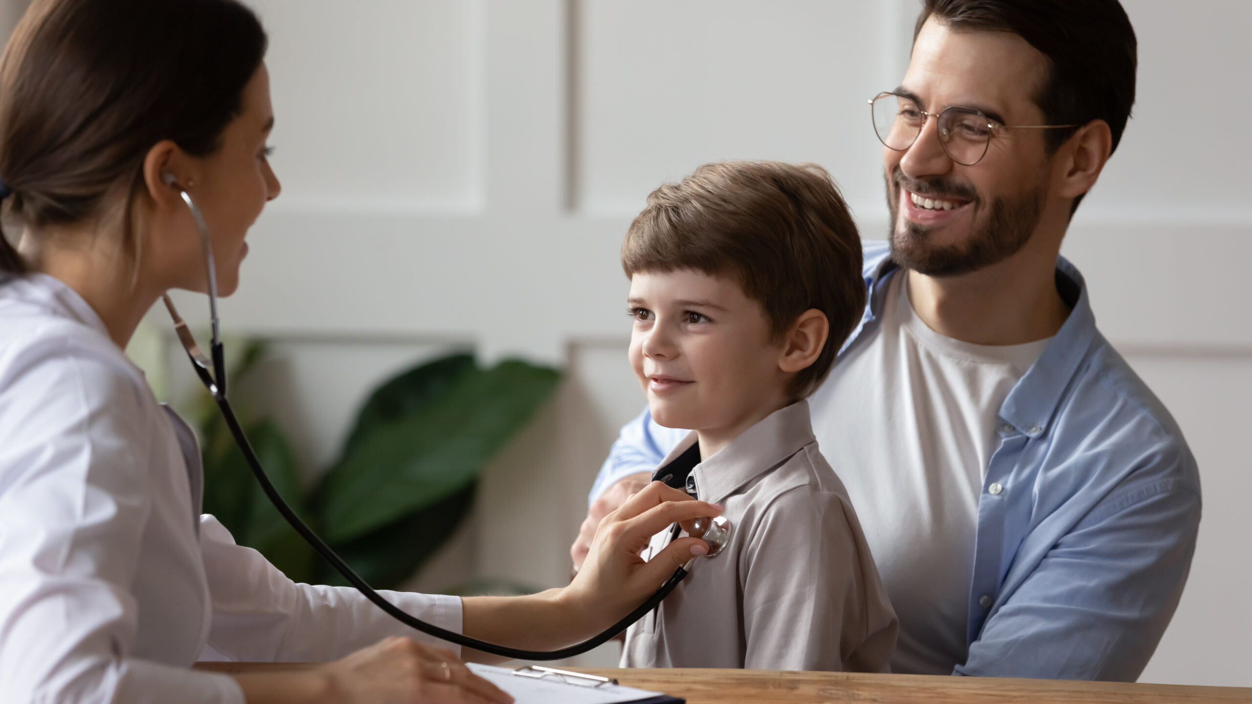 Doctor listening to child's chest through stethoscope while child sits on father's lap